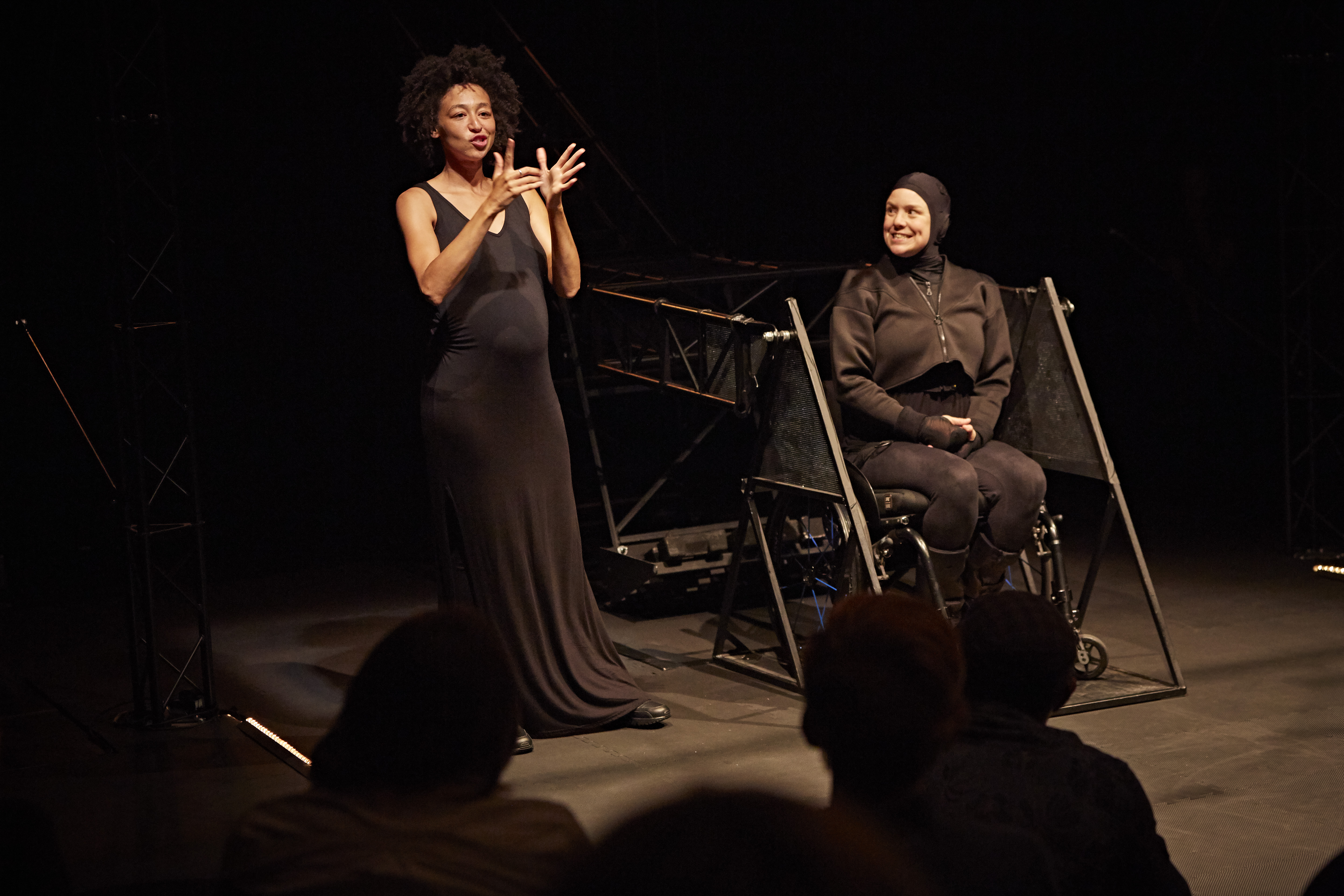 Intro "Not I" Intro der Performance "Not I". Links spricht die Gebärdensprachdolmetscherin. Rechts sitzt Jess in ihrem Rollstuhl auf der Heberampe, die sie in der Performance in die Höhe hebt. Jess ist ganz in schwarz gekleidet, auch ihr Kopf ist von einer schwarzen Kapuze bedeckt, sodass man nur ihr Gesicht sieht.