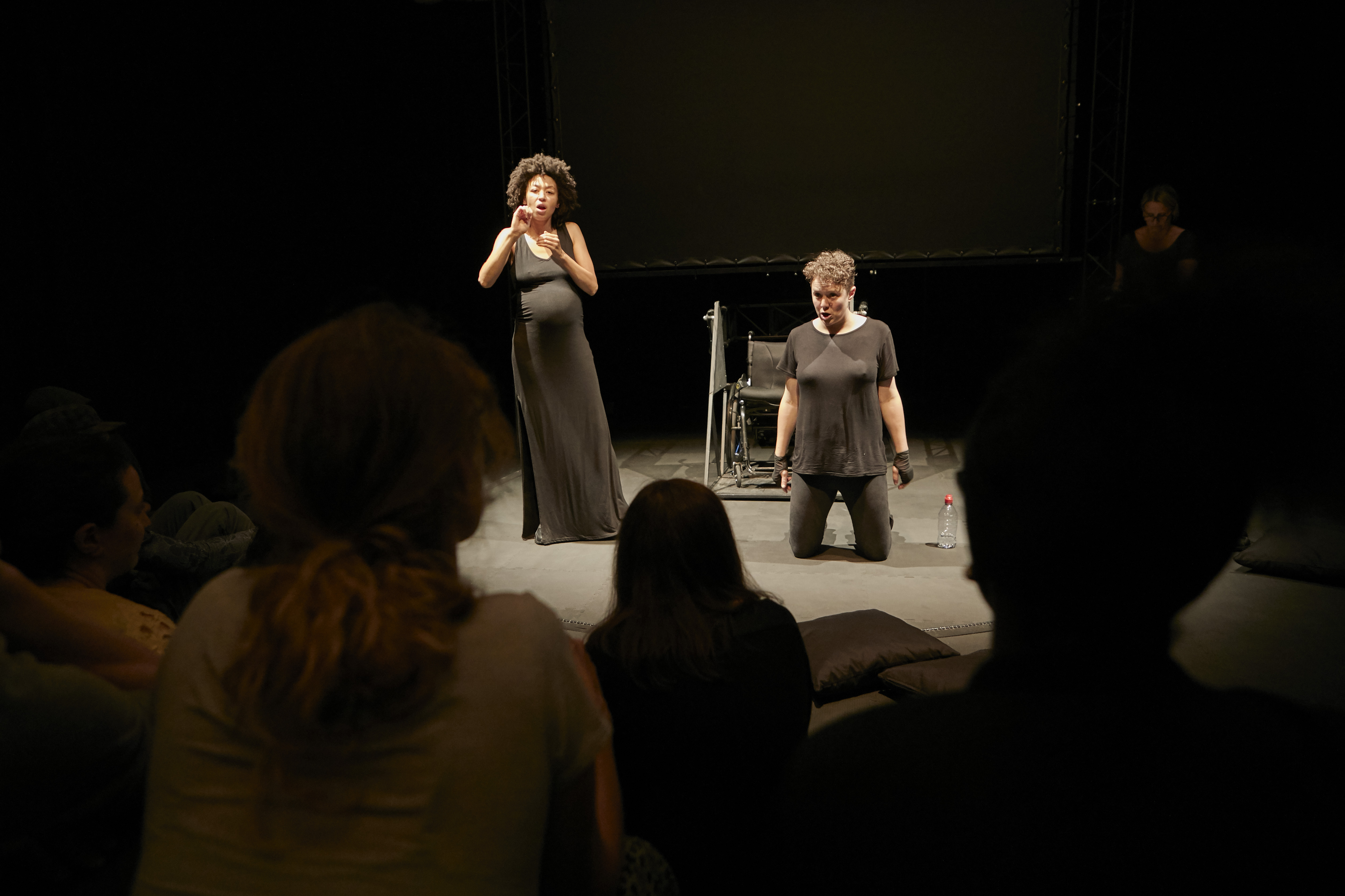 Diskussion nach der Performance von "Not I" Diskussion nach der Performance von "Not I". Der Blick der Kamera geht durch das Publikum auf die Bühne. Links steht die Gebärdensprachdolmetscherin, rechts kniet Jess Thom vor ihrem Rollstuhl. Beide sind in Schwarz gekleidet.