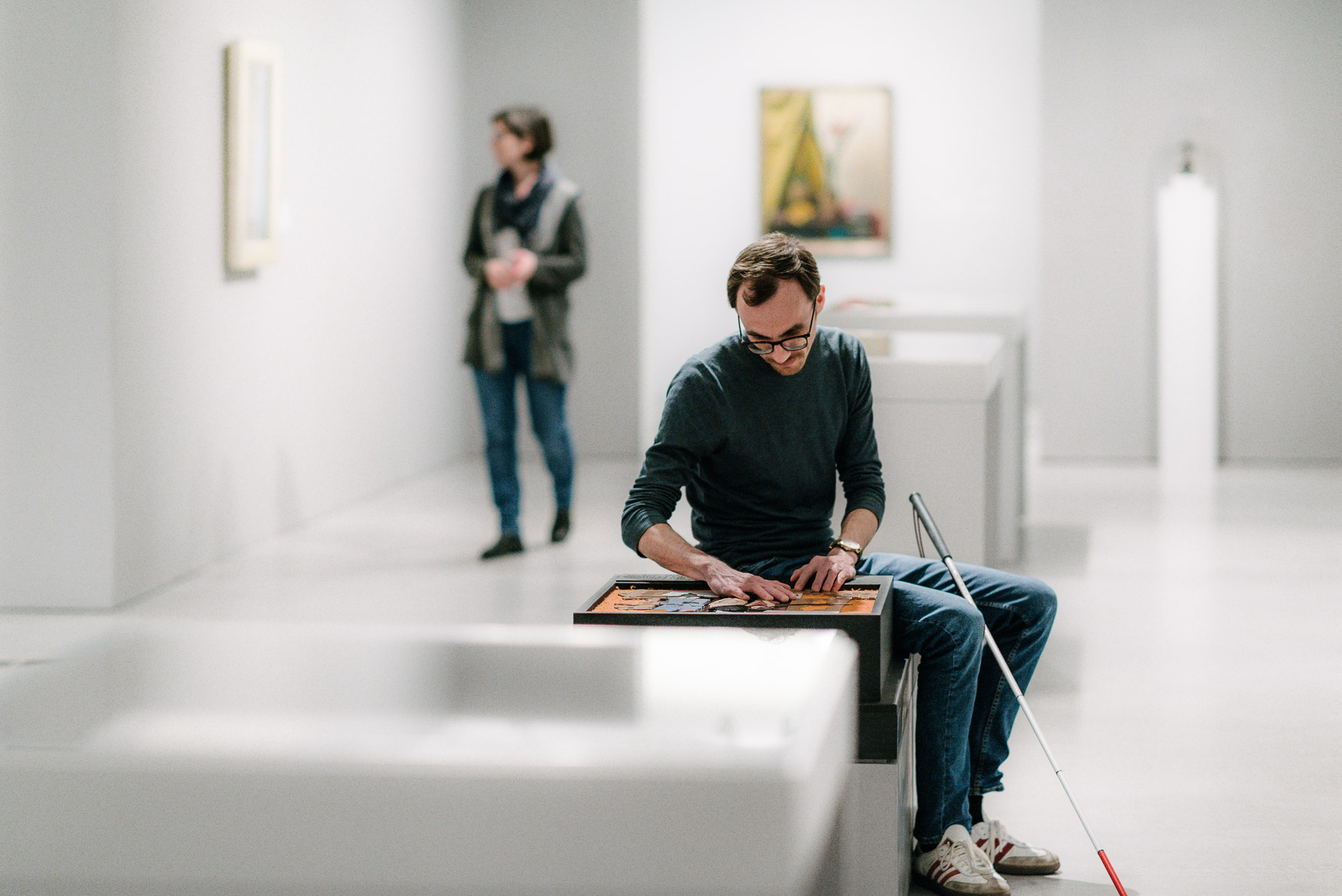 Kunst zum Anfassen in der Berlinischen Galerie. Andreas Krüger sitzt in einem Ausstellungsraum, im Hintergrund sind Bilder aufgehängt, vor einem der Bilder steht eine Besucherin. Vor Andreas Krüger befindet sich ein Tastmodell, das er mit den Händen ertastet, ein Langstock lehnt an seinem Knie.