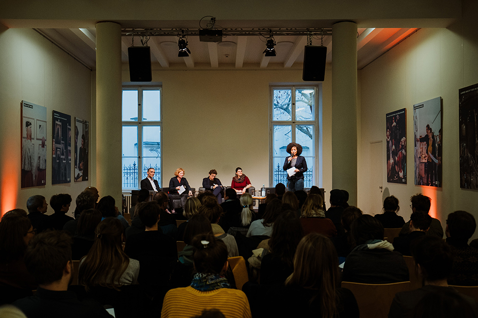 Podiumsdiskussion bei der Jahrestagung des LMB 2019. Murat Akan (erster von links) Blick von hinten auf einen vollen Saal. Auf dem Podium sitzen vier Referent*innen, eine Moderatorin mit einem Mikro in der Hand steht daneben.