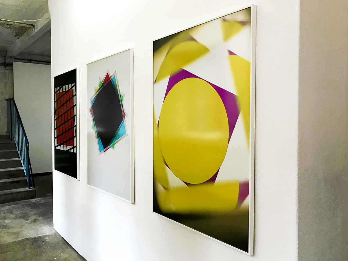 ASPEKTE KONKRET Man sieht drei Bilder an einer weißen Wand hängen, wobei zwei der Bilder zu weit entfernt vom Betrachter sind. Das dritte Bild mit dem Titel Dichroitisch zeigt geometrische Formen in gelb und lila.