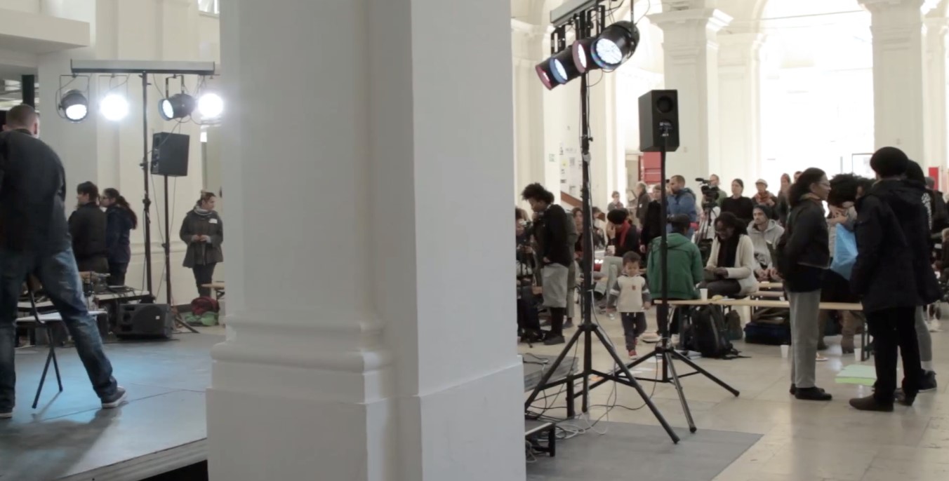 Standbild Konferenz Vernetzt euch Blick auf ein Publikum in einer repräsentativen Halle, eine Bühne und Scheinwerfer sind an der Seite zu sehen