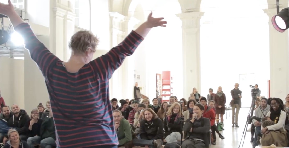 Standbild Konferenz Vernetzt euch Blick von der Bühne aus auf das Publikum. Eine Performerin ist von hinten zu sehen, sie streckt die Arme in die Höhe. Sie trägt ein geringeltes Hemd und ein Hörgerät.