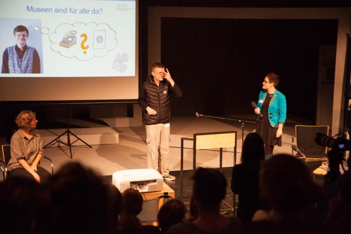 Prästentation "Nueva - Menschen mit Behinderungen evaluieren" mit Patrick Marx Auf der Bühne gebärdet Patrick Marx, hinter ihm seine Powerpointpräsentation