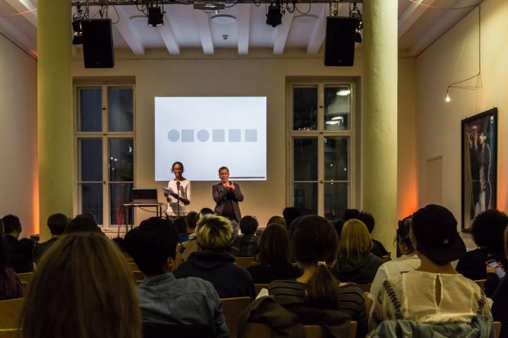 Die Autorin Sharon Dodua Otoo steht auf der Bühne und liest. Neben ihr steht ein DGS-Dolmetscher. Vor der Bühne sitzen Menschen und hören zu.