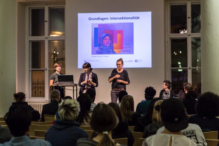 Auf der Bühne stehen Mitarbeitende von Diversity Arts Culture und präsentieren ihre Arbeit. Im Hintergrund gibt es eine Powerpointpräsentation, auf der man ein Bild zu &quot;Intersektionalität&quot; sieht. Vor der Bühne sitzen Menschen und hören zu.