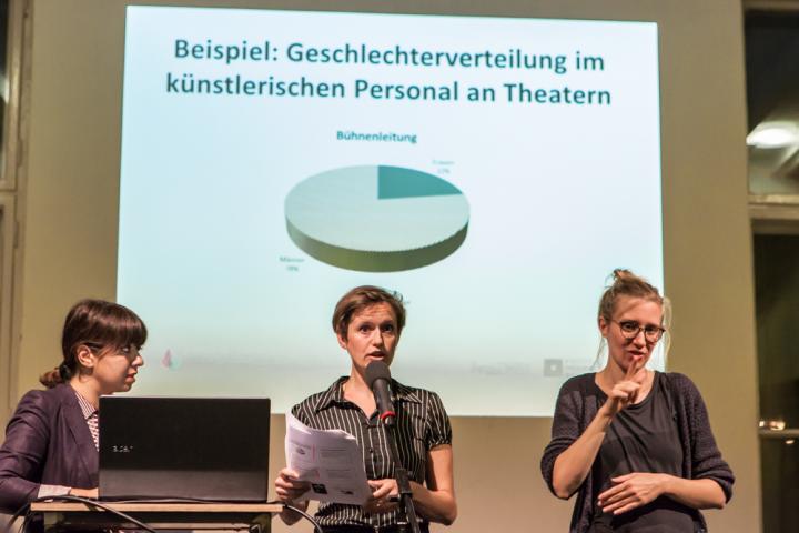 Im Hintergrund: Powerpointfolie mit einem Tortendiagramm zur Geschlechter-Verteilung am Theater. Im Vordergrund zwei Mitarbeitende von Diversity Arts Culture und eine DGS-Dolmetscherin.
