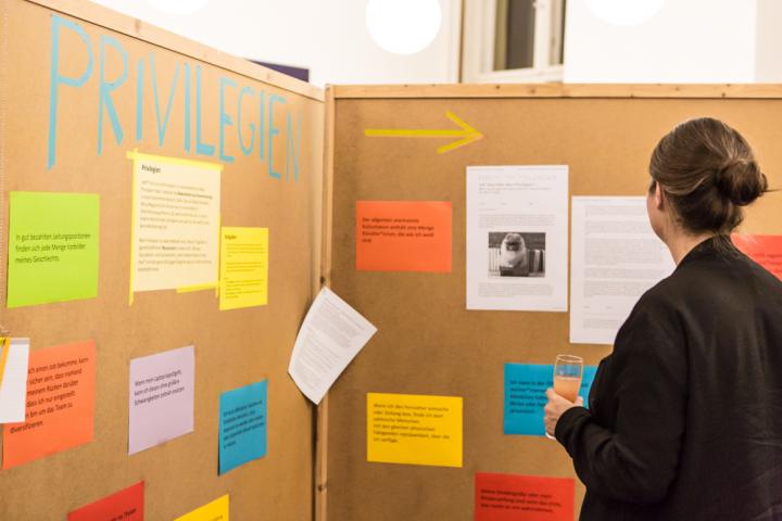 Eine Frau betrachtet Informationen auf Stellwänden. Auf der einen Stellwand steht groß Privilegien, darunter hängen viele bunte Zettel.