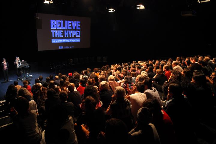 Im Hintergrund sieht man die Bühne, vorne eine Tribühne, die bis auf den letzten Platz besetzt ist