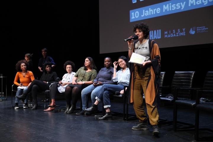 Die Moderatorin hält ein Mikrofon in der Hand, links neben ihr sitzen die Sprecherinnen der eingeladenen Initiativen