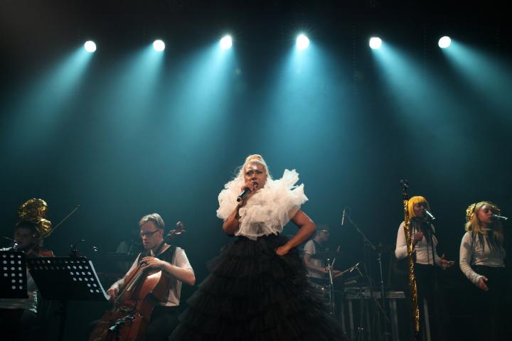 Die Sängerin Ebony Bones steht mit einem Mikrofon in der Hand auf der Bühne. Links neben ihr sitzt ein Cellist, rechts neben ihr stehen zwei Sängerinnen mit gelben Perrücken. Die Bühne ist in ein türkisenes Licht getaucht.