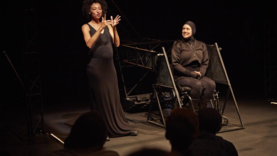 Intro "Not I" Intro der Performance "Not I". Links spricht die Gebärdensprachdolmetscherin. Rechts sitzt Jess in ihrem Rollstuhl auf der Heberampe, die sie in der Performance in die Höhe hebt. Jess ist ganz in schwarz gekleidet, auch ihr Kopf ist von einer schwarzen Kapuze bedeckt, sodass man nur ihr Gesicht sieht.