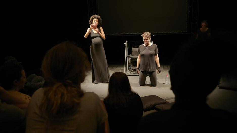 Diskussion nach der Performance von "Not I" Diskussion nach der Performance von "Not I". Der Blick der Kamera geht durch das Publikum auf die Bühne. Links steht die Gebärdensprachdolmetscherin, rechts kniet Jess Thom vor ihrem Rollstuhl. Beide sind in Schwarz gekleidet.