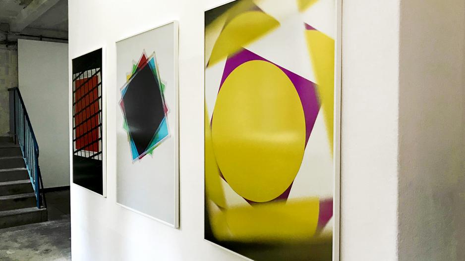 ASPEKTE KONKRET Man sieht drei Bilder an einer weißen Wand hängen, wobei zwei der Bilder zu weit entfernt vom Betrachter sind. Das dritte Bild mit dem Titel Dichroitisch zeigt geometrische Formen in gelb und lila.