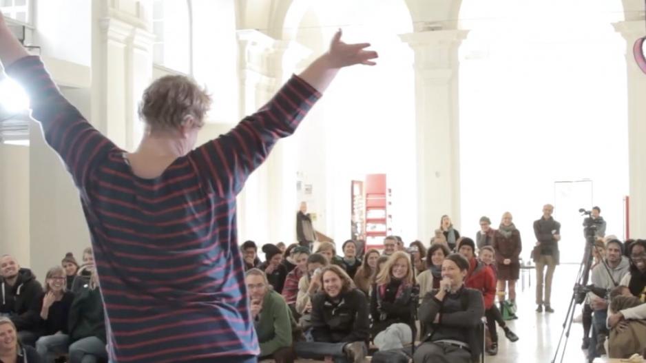 Standbild Konferenz Vernetzt euch Blick von der Bühne aus auf das Publikum. Eine Performerin ist von hinten zu sehen, sie streckt die Arme in die Höhe. Sie trägt ein geringeltes Hemd und ein Hörgerät.